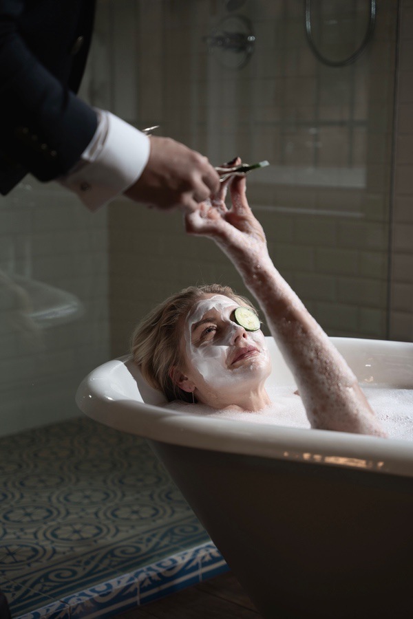 Ein Gast entspannt mit Gesichtsmaske in der Badewanne, der Butler assistiert
